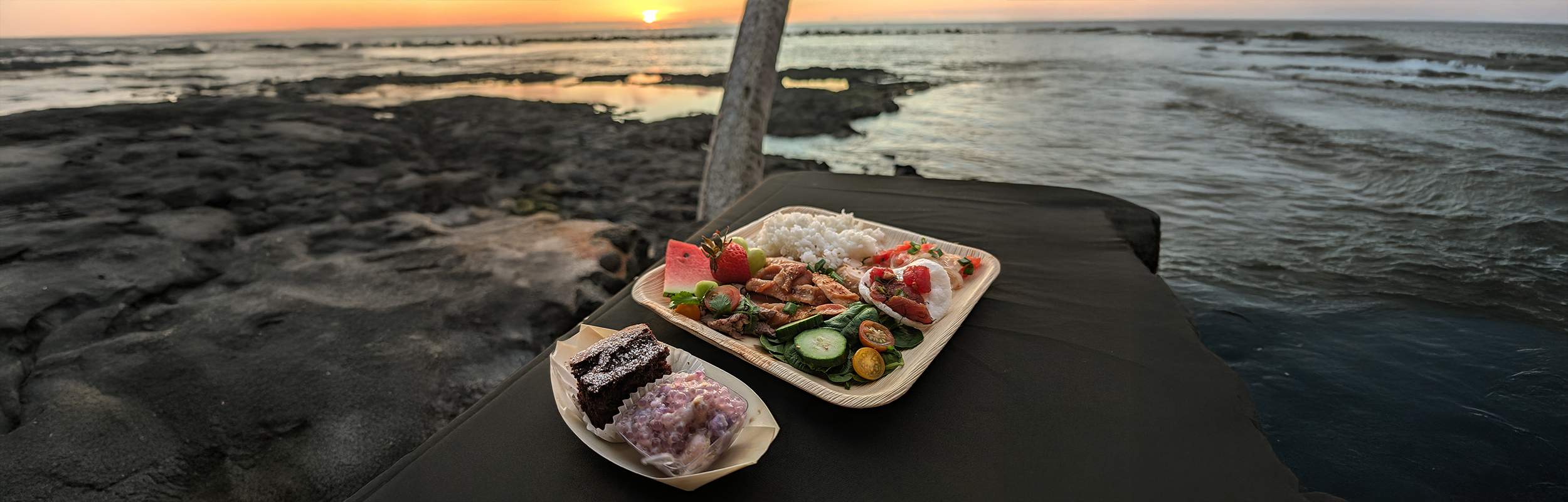 A dinner plate of rice, grilled meat, salad, fruit and dessert on a table by the lava-rock shoreline at sunset
